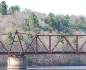 The railroad swing bridge in spring, with leafless hardwoods and dark green White pines on the bluff behind it.